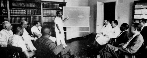 Black man standing in front of a classroom of other black men, pointing at a chalkboard